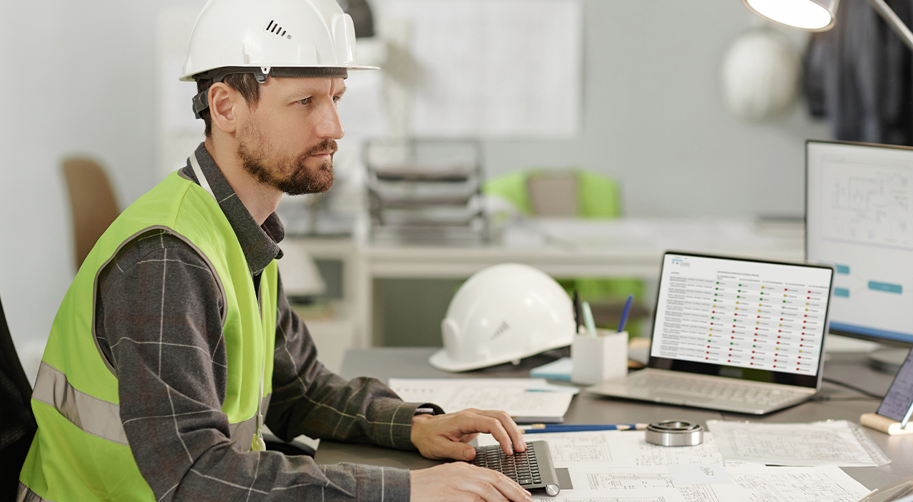 A construction professional in a hard hat and safety vest uses a laptop displaying a Trimble project management dashboard.