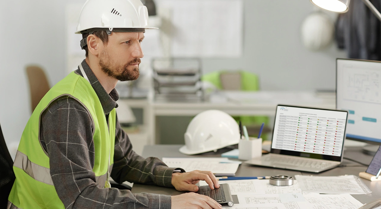 A construction professional in a hard hat and safety vest uses a laptop displaying a Trimble project management dashboard.