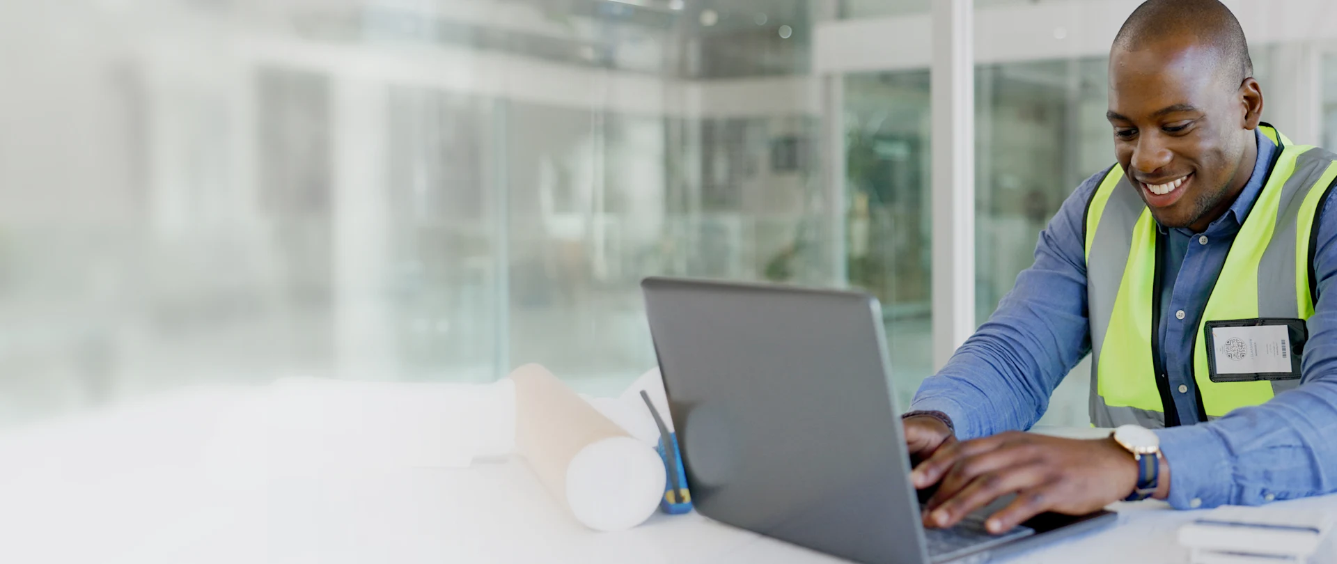 A smiling Black professional in a high-visibility vest works on a laptop with rolled blueprints and a QR code badge in a modern construction office.
