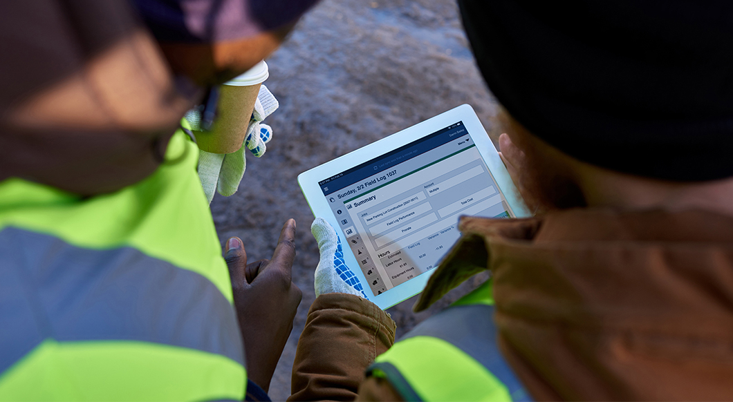 Two construction workers review a field log summary on a tablet using Trimble software at a job site.