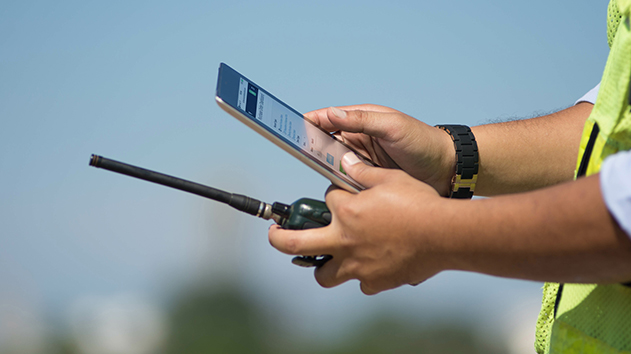 A worker in a high-visibility vest holds a walkie-talkie and a mobile device displaying the Trimble ProjectSight project dashboard.