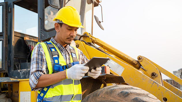 A construction worker in a yellow hard hat and safety vest reviews data on a rugged tablet in front of a yellow front-end loader on a job site.