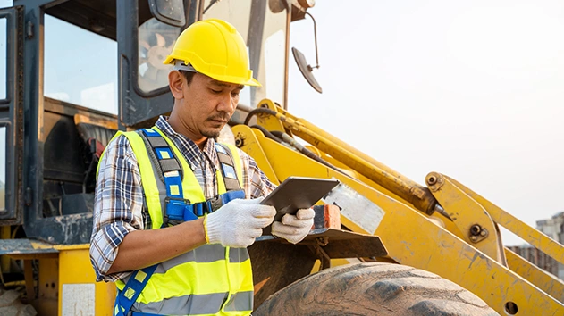 A construction worker in a yellow hard hat and safety vest reviews data on a rugged tablet in front of a yellow front-end loader on a job site.