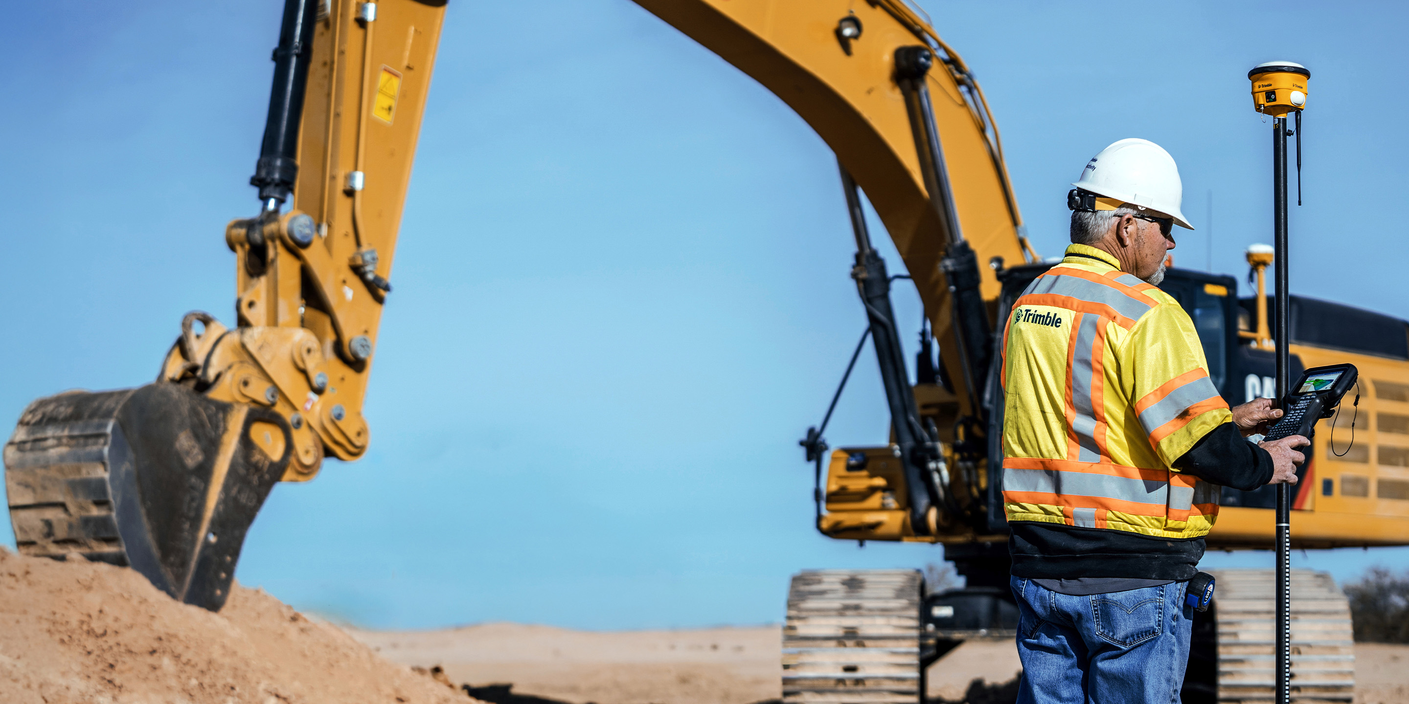 A worker in a safety vest uses a handheld Trimble controller at a construction site with a large yellow excavator in the background.