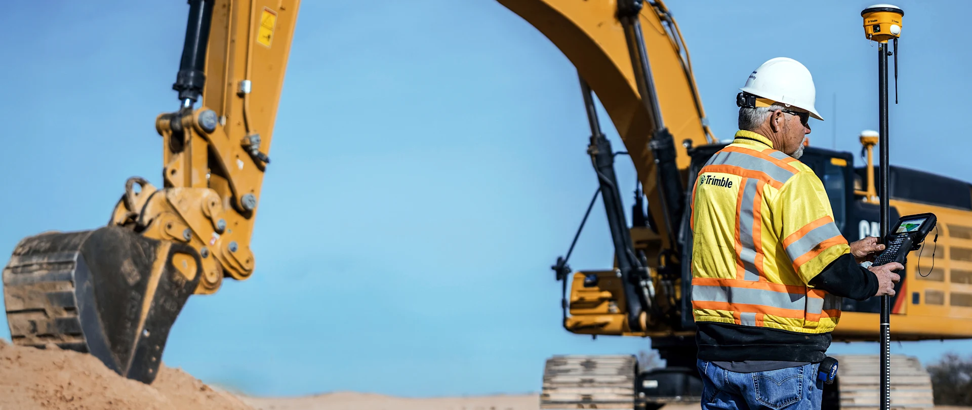 A worker in a safety vest uses a handheld Trimble controller at a construction site with a large yellow excavator in the background.