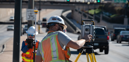 Two surveyors use a Trimble scanning total station and GNSS rover at a highway construction site with traffic in the background.