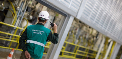 A worker in a green vest and white hard hat, labeled "OPERAÇÃO," communicates via radio in an industrial setting with yellow railings and machinery.