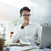 A man wearing glasses sits at a desk with a laptop, looking thoughtful.