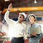 Two people in hard hats discuss work in an industrial plant; a man points upward while a woman holding a laptop looks in the same direction.