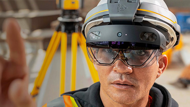 A close-up shot of a man wearing a white hard hat with a gray and yellow stripe, and a pair of futuristic-looking augmented reality glasses.