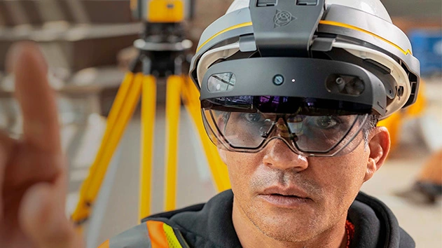 A close-up shot of a man wearing a white hard hat with a gray and yellow stripe, and a pair of futuristic-looking augmented reality glasses.