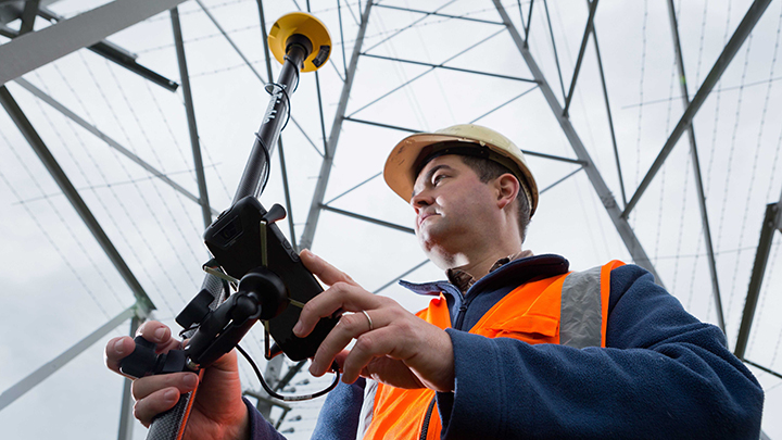 2016: Site engineer with hard hat and reflective vest uses Trimble Catalyst under utility pylon
