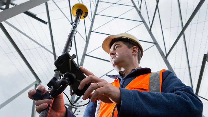 2016: Site engineer with hard hat and reflective vest uses Trimble Catalyst under utility pylon