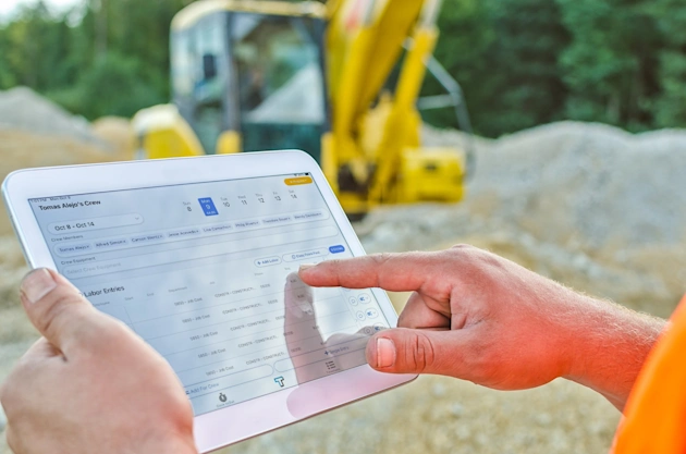 A person uses Trimble labor entry software on a tablet at a construction site with a blurred yellow excavator in the background.
