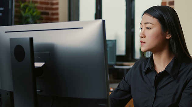 A woman with long dark hair and a black collared shirt looks intently at a computer monitor in an office with brick walls.