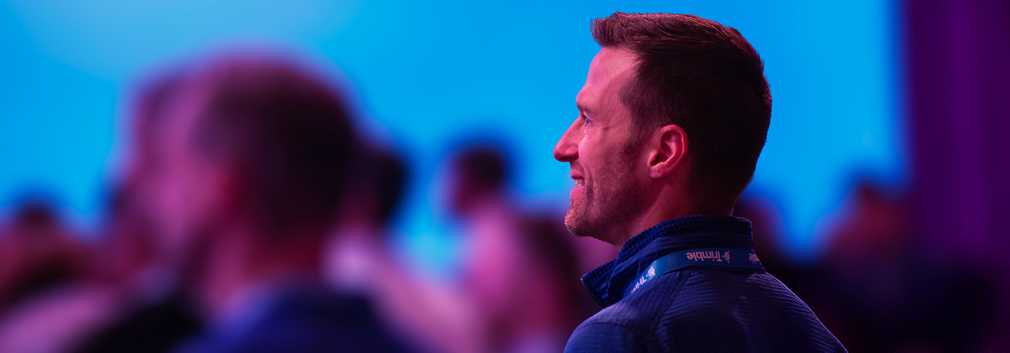 A smiling man in profile wearing a Trimble lanyard at a corporate event, with a blurred background of blue and purple stage lighting.