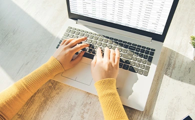 Overhead view of a person in a yellow sweater typing on a laptop displaying a spreadsheet on a light wooden desk in natural sunlight.