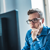 A focused man in glasses and a blue shirt rests his chin on his hand while looking at a computer monitor.