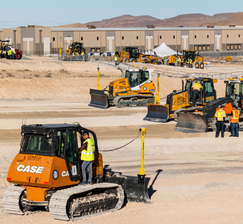 An orange CASE bulldozer with an operator stands on a dirt construction site in the foreground of Offsite Expo.jpg. Other heavy machinery, workers, and a large building are visible in the background under a clear sky.