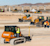 An orange CASE bulldozer with an operator stands on a dirt construction site in the foreground of Offsite Expo.jpg. Other heavy machinery, workers, and a large building are visible in the background under a clear sky.
