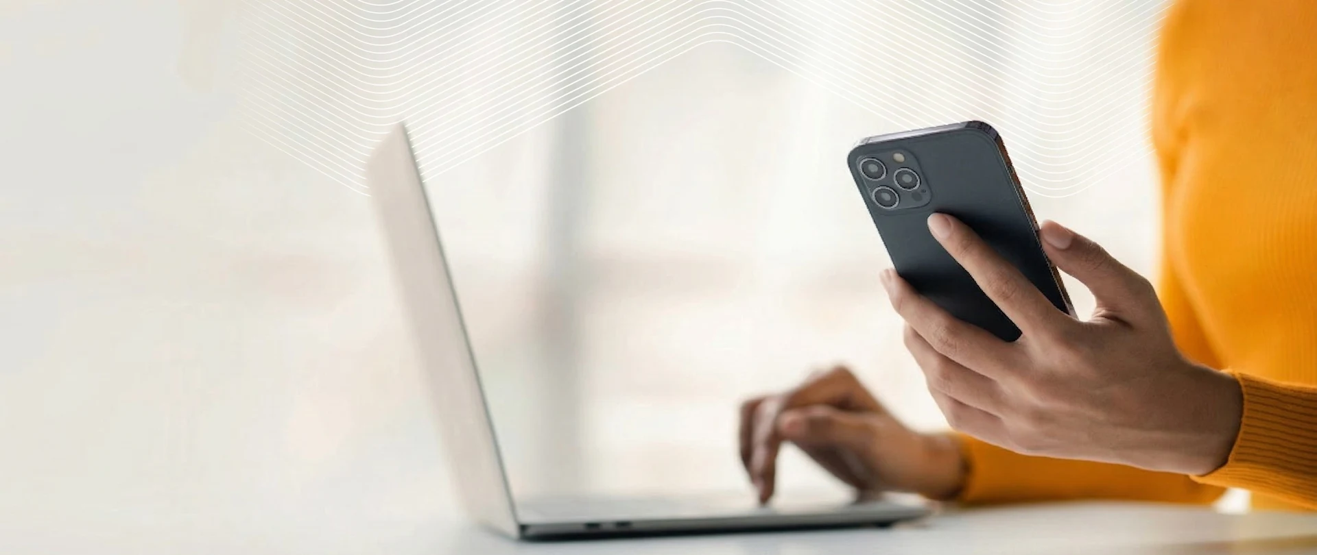 Close-up of a person in a yellow sweater holding a smartphone while their other hand uses a laptop trackpad.