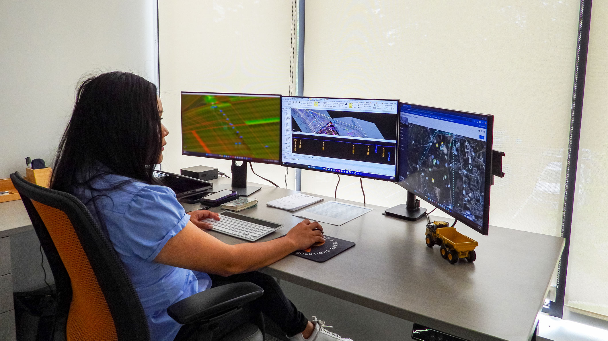 A professional woman works with Trimble Business Center and site management software on triple monitors at a desk with a yellow toy dump truck.
