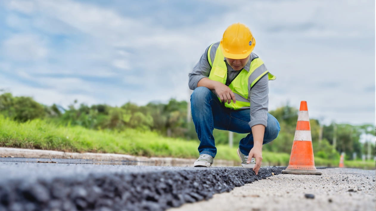 Construction worker in a yellow hard hat and safety vest inspecting fresh asphalt on a new road.