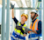 Two construction workers in safety gear inspect overhead industrial pipes using digital tablets.