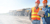 Two construction professionals wearing hard hats and safety vests stand on a jobsite reviewing data on a tablet. In the background, a line of large yellow haul trucks is parked along a dirt road under a bright, clear sky.