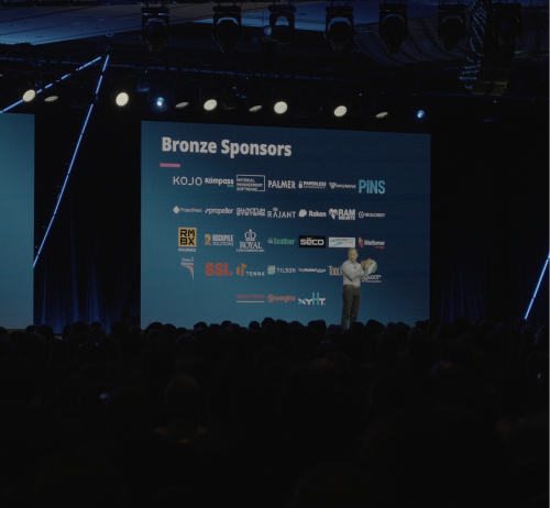An auditorium stage screen titled "Bronze Sponsors" displays multiple rows of company logos. A presenter stands to the right of the screen in front of a large, seated audience in a dimly lit conference hall.