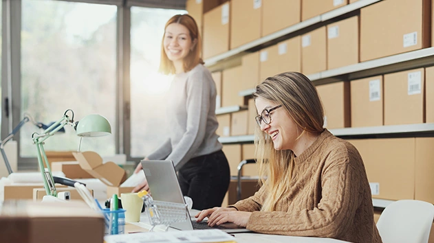 Two women smile in a warehouse. One sits at a desk using a laptop while the other stands near shelves filled with cardboard shipping boxes.