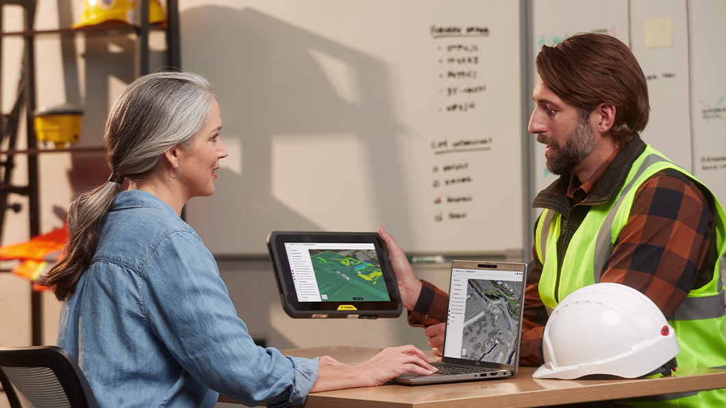Two professionals discuss site data using a laptop and a rugged tablet in an office with construction gear in the background.