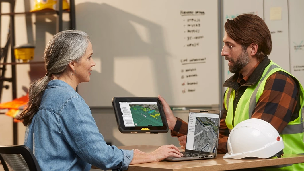 Two professionals discuss site data using a laptop and a rugged tablet in an office with construction gear in the background.