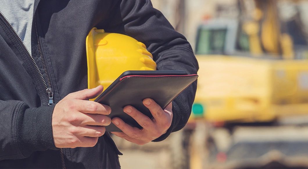 A person in construction gear holds a yellow hard hat and a Trimble rugged tablet on a job site with a yellow excavator in the background.