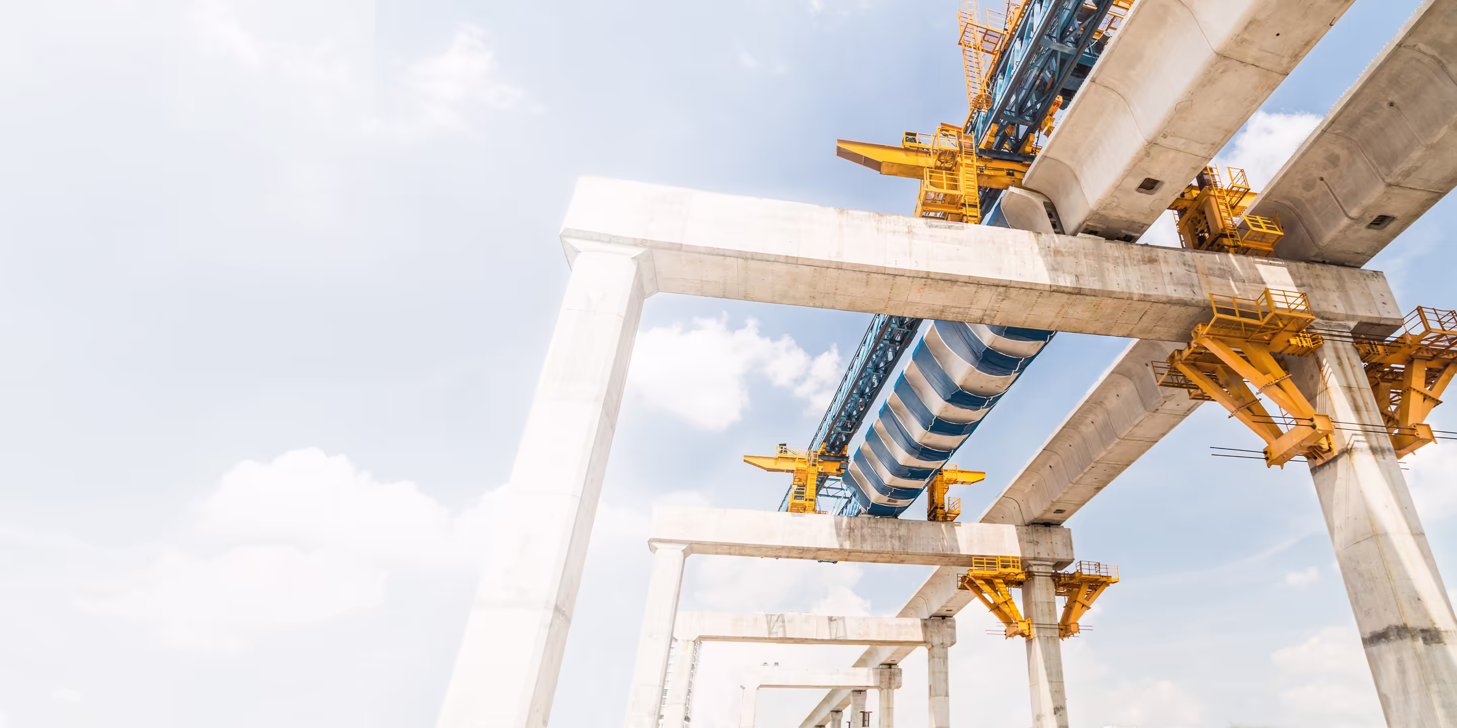 A blue and white striped launching gantry machine constructs an elevated concrete bridge between piers against a bright sky.