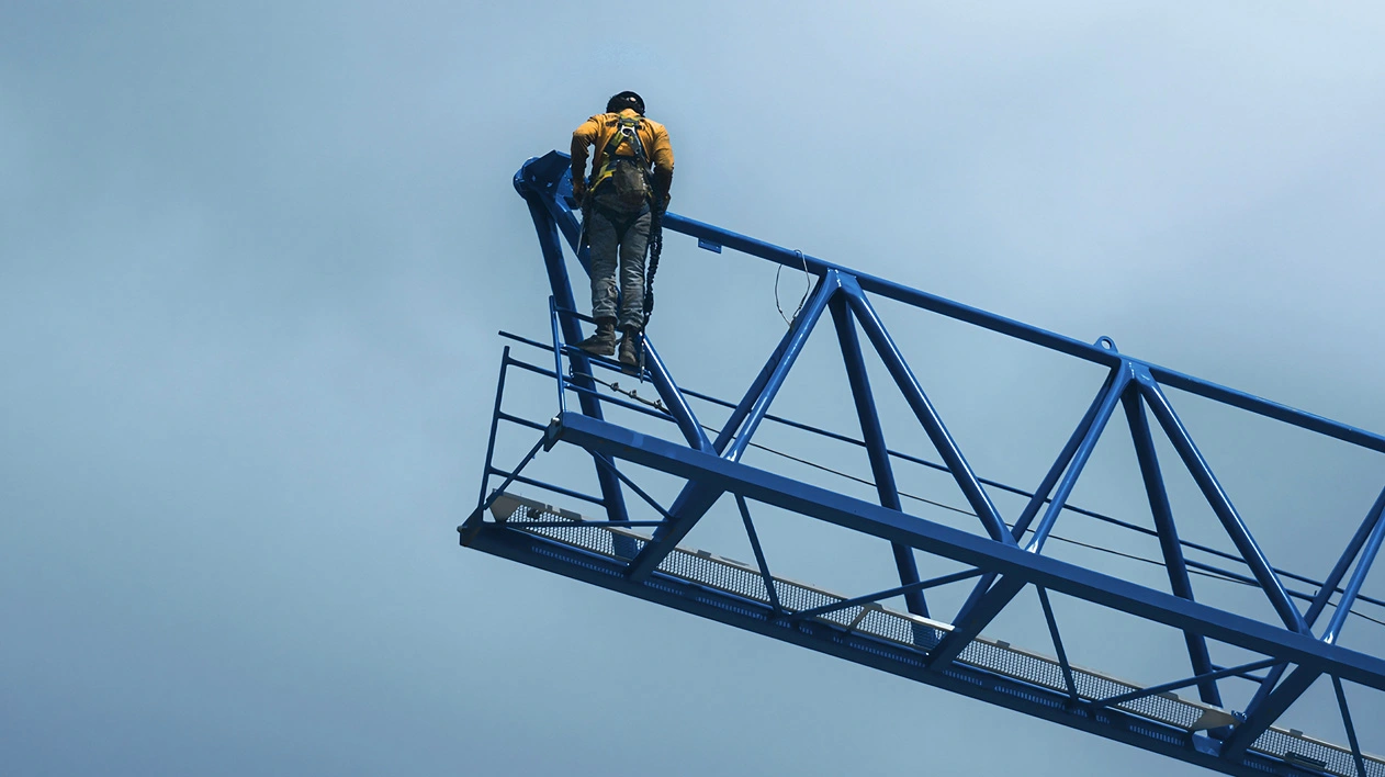 A construction worker in a safety harness stands on a blue lattice crane jib against a clear sky.