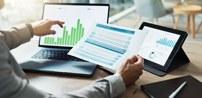 A person analyzes green bar graphs on a laptop and printed spreadsheet, with a tablet showing a line graph nearby, conveying a data-focused workspace.