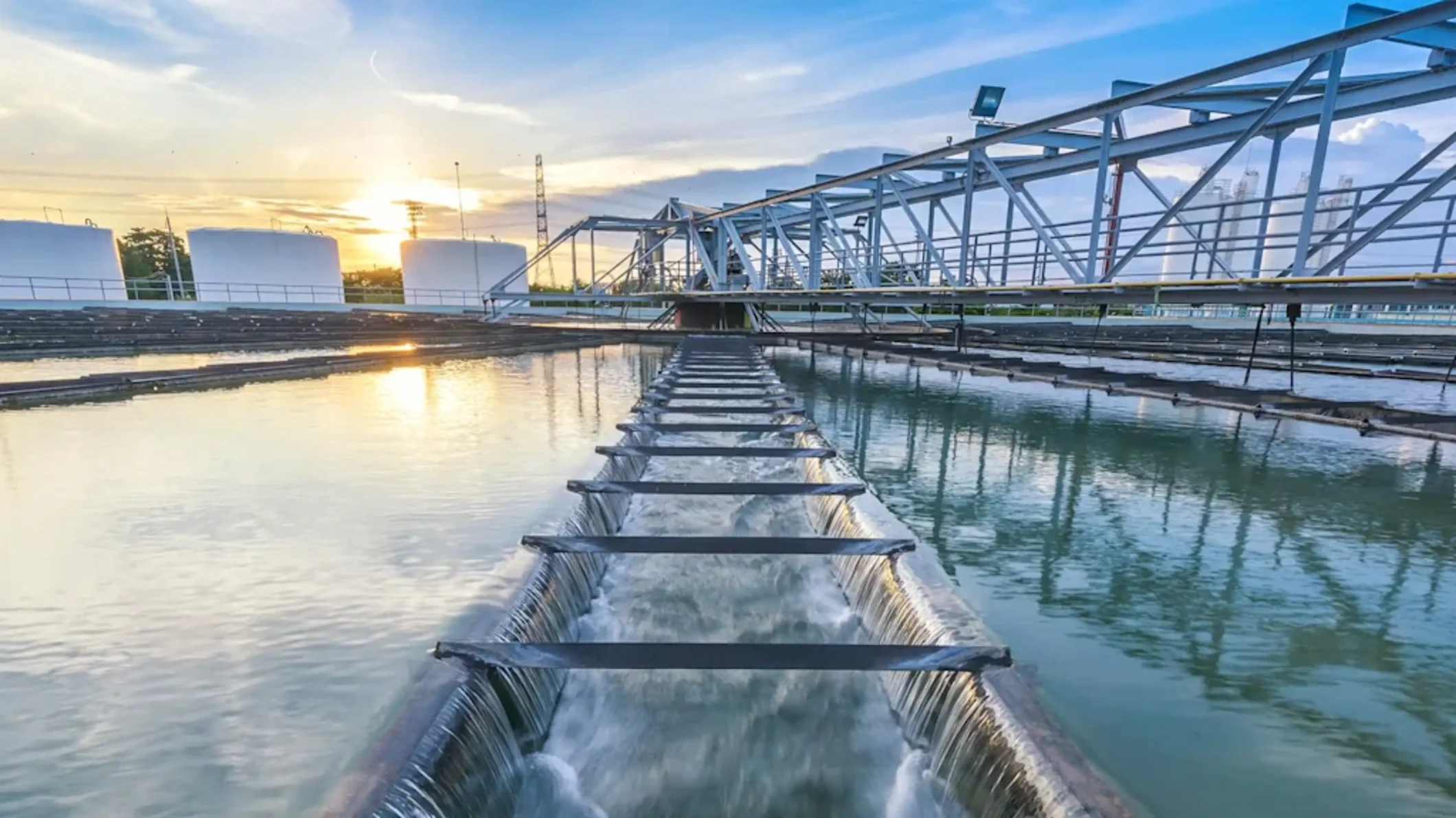 Active water treatment facility: Water flowing over multiple weirs in clarifier basins. A steel truss bridge and large white industrial storage tanks are visible under a vibrant sunset sky.
