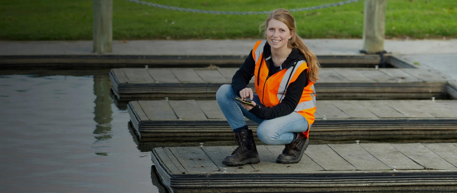 Woman in reflective vest and boots on dock smiling at camera using mobile device