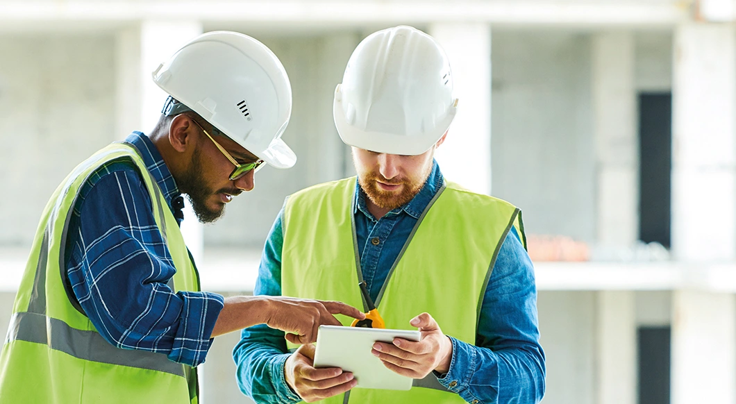Two construction workers in hard hats and safety vests review project plans on a tablet at a building site.