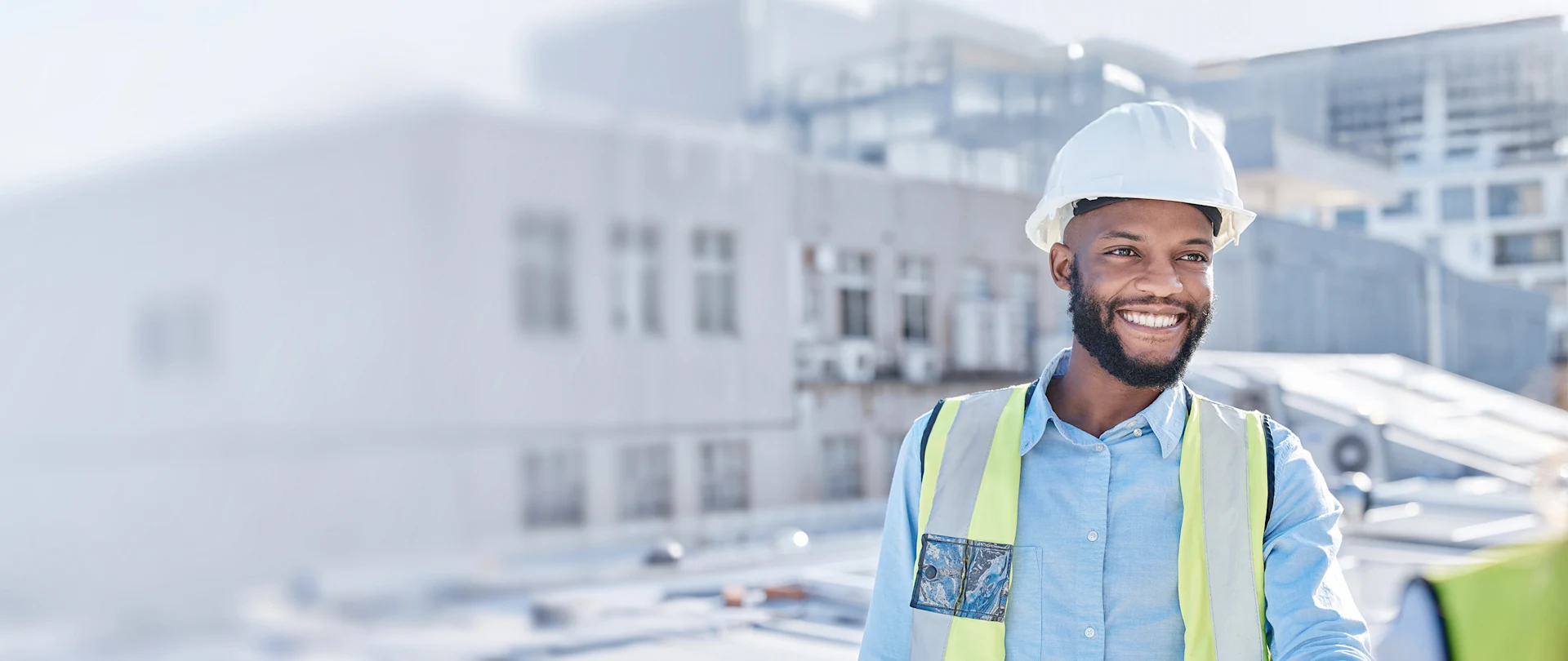 A smiling African American construction worker in a white hard hat and yellow safety vest stands on a rooftop construction site.