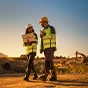 Two construction workers in safety gear walk across a site at sunset, one holding a Trimble rugged tablet.