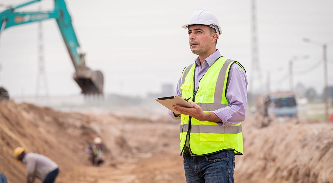 A construction professional uses a Trimble rugged tablet at a jobsite with a turquoise excavator in the background.