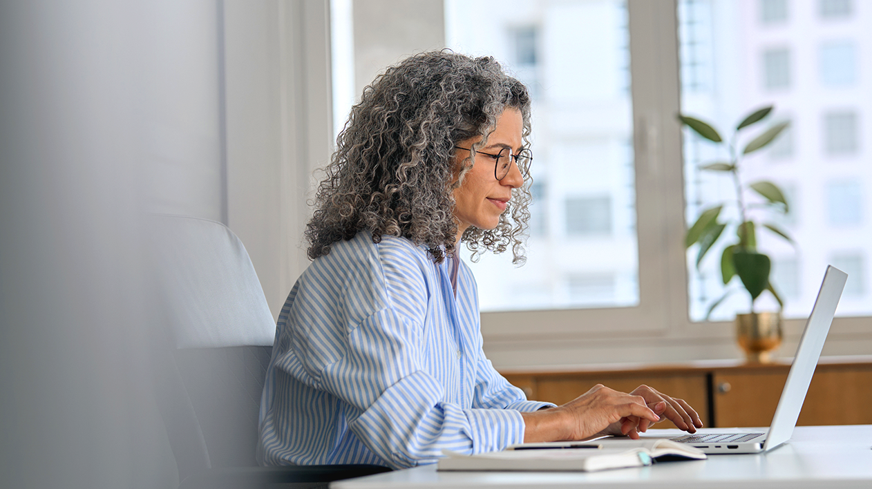 A professional woman with curly gray hair and glasses working on a laptop at a white desk in a bright, modern office.