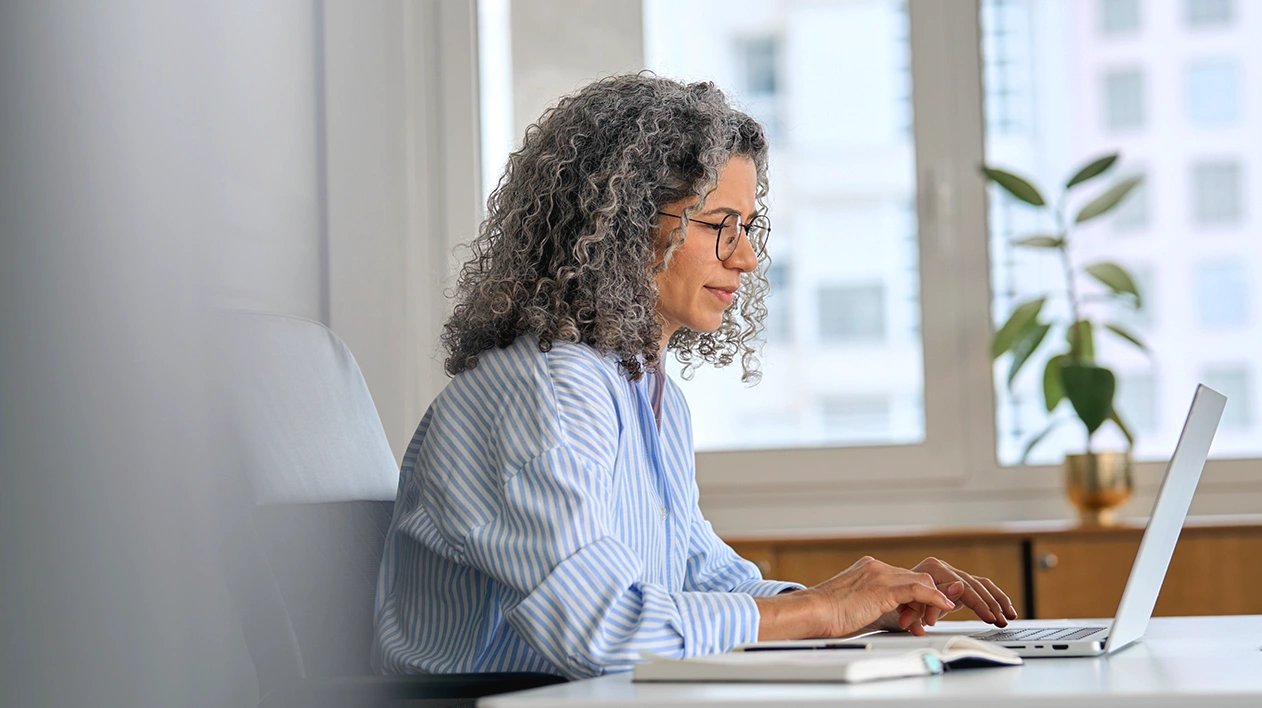 A professional woman with curly gray hair and glasses working on a laptop at a white desk in a bright, modern office.