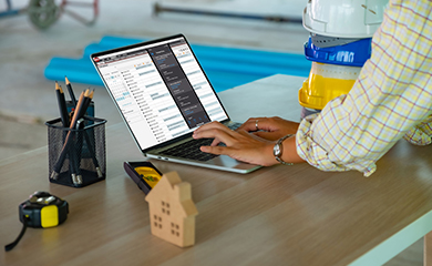 A construction professional using Trimble ProjectSight software on a laptop at a job site, with stacked hard hats and tools on a desk.