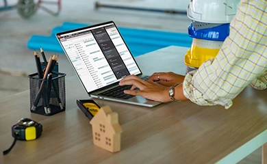 A construction professional using Trimble ProjectSight software on a laptop at a job site, with stacked hard hats and tools on a desk.
