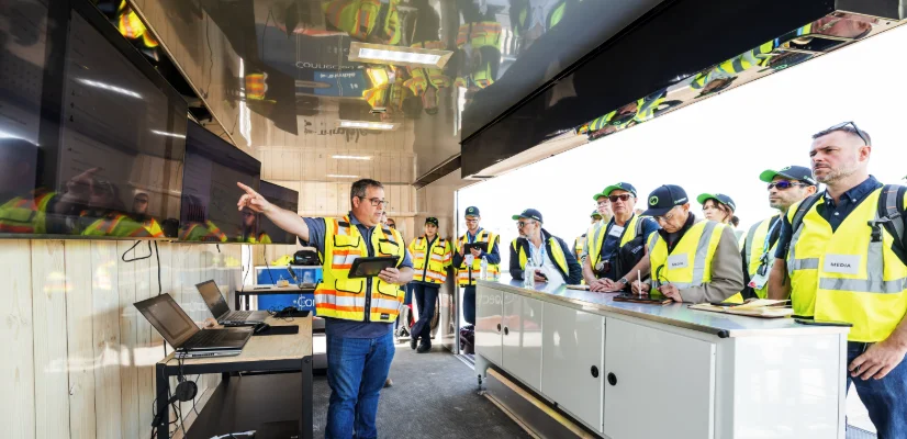 An instructor at the Trimble Dimensions offsite points to a large screen while speaking to a group of focused attendees in PPE