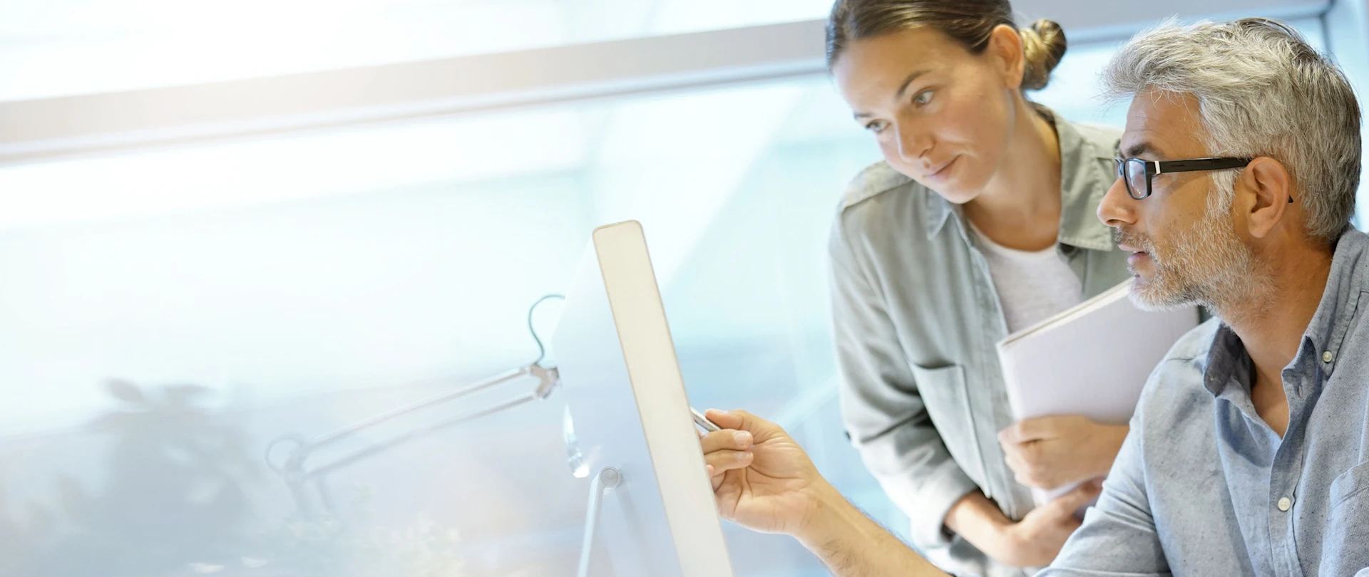 Two office colleagues collaborate at a computer; a man points at the screen while a woman stands nearby holding documents.