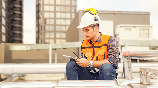 A construction worker on a rooftop using a rugged tablet and radio, wearing a white hard hat, yellow ear muffs, and an orange safety vest.